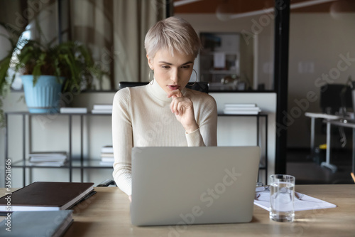 Concentrated businesswoman sitting at desk in contemporary office room looking at laptop screen thinking over problem solution, typing response to client corporate customer, busy working day concept