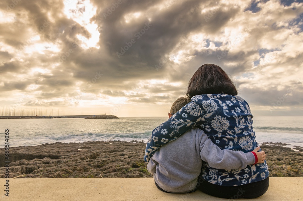 Mom with son sitting and hugging each other on the wall facing the sea