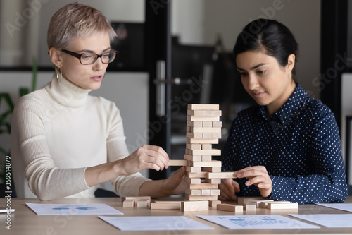 Caucasian Indian two businesswomen sit at desk in office build tower of wooden blocks involved in game play. Team building activity, teamwork, business strategizing, strategic thinking concept