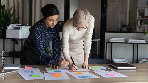 Multiracial businesswomen work on project statistics, prepare financial report use post-it memo notepapers making notes do sales overview, colleagues analysing charts data, teamwork brainstorm concept