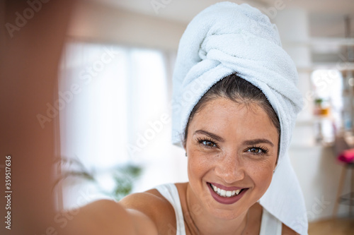 Self portrait of charming, stylish, pretty, attractive model after bath wrapped in towel shooting selfie on front camera, enjoying procedure. Young woman in her apartment doing her beauty care routine