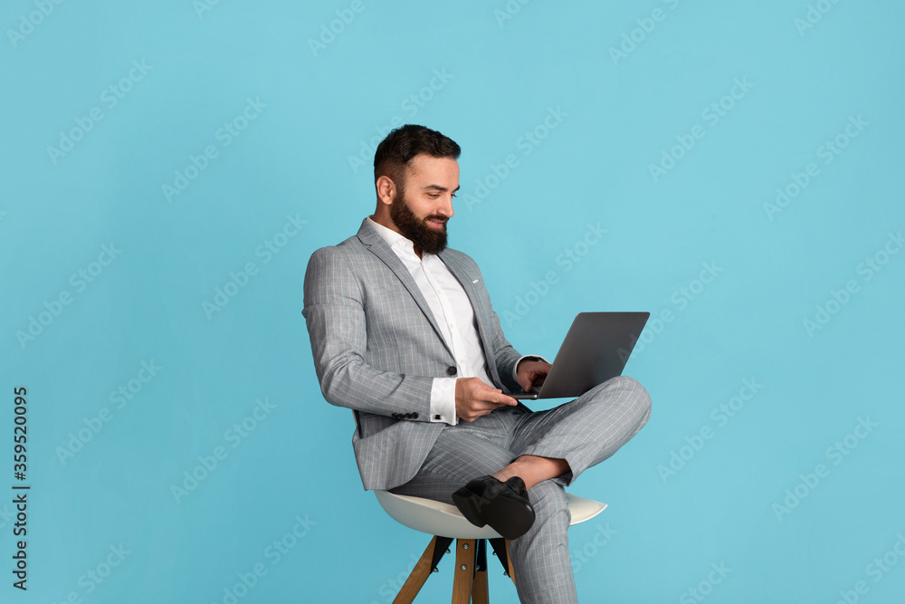 Attractive businessman using laptop computer while sitting on chair against blue background Attractive businessman using laptop computer while sitting on chair against blue background