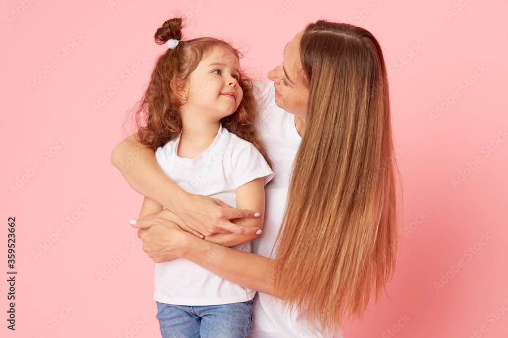 Mom and daughter in white t-shirts and jeans play and hug on a pink background. Caring for loved ones. Happy motherhood Mom and daughter in white t-shirts and jeans play and hug on a pink background. Caring for loved ones. Happy motherhood