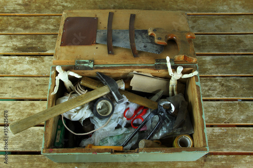 Overhead shot of old toolbox. On view: saw, hammers, pliers, adhesive tape, scissors and two wooden mannequins. On wooden table.