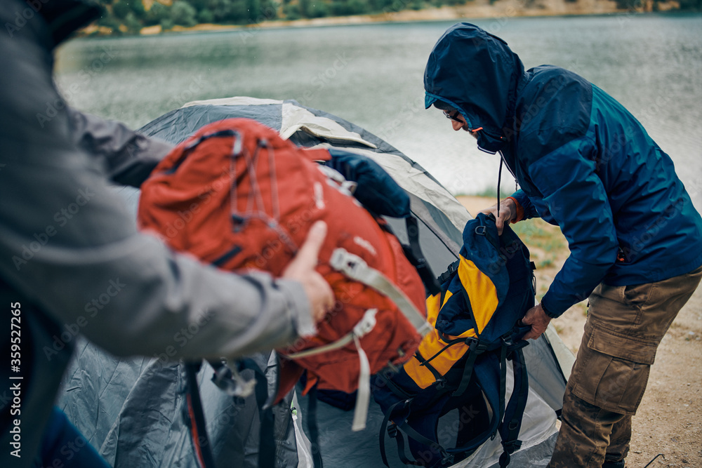 Fototapeta premium Two men on camping trip putting backpacks into tent by the lake on rainy and cold weather.
