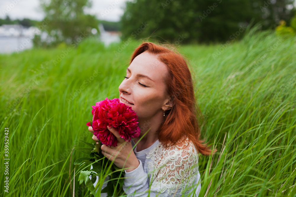 Fototapeta premium Smiling redhead girl with peonies in grass. Profile shot