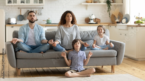 Relaxed young family with cute children sitting and meditating with closed eyes. Attractive wife and husband with son and daughter do breathing exercises. Parents with kids doing yoga together. photo