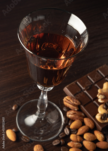 Cognac wine glass with amber cognac, nuts lying near the wine glass, coffe grains, chocolate bar on a dark background.