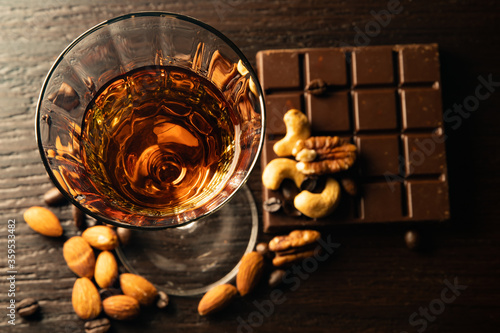 Cognac wine glass with amber cognac, nuts lying near the wine glass, coffe grains, chocolate bar on a dark background. View from the top.