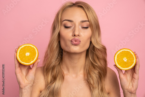 Attractive comic positive nude girl with beaming smile having two pieces of orange sending blowing kiss with pout lips, close eyes, isolated on pink background