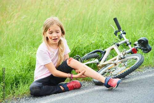 Sad crying little child girl fell from the bike in the summer park. Bleeding on hands and feet. Bicycle accident. Injuries while cycling. Accidents and injuries to children during summer activities