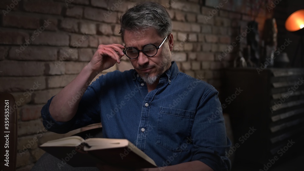 Mature man in his 50s reading book at home sitting in dark living room. Mature man in his 50s reading book at home sitting in dark living room.