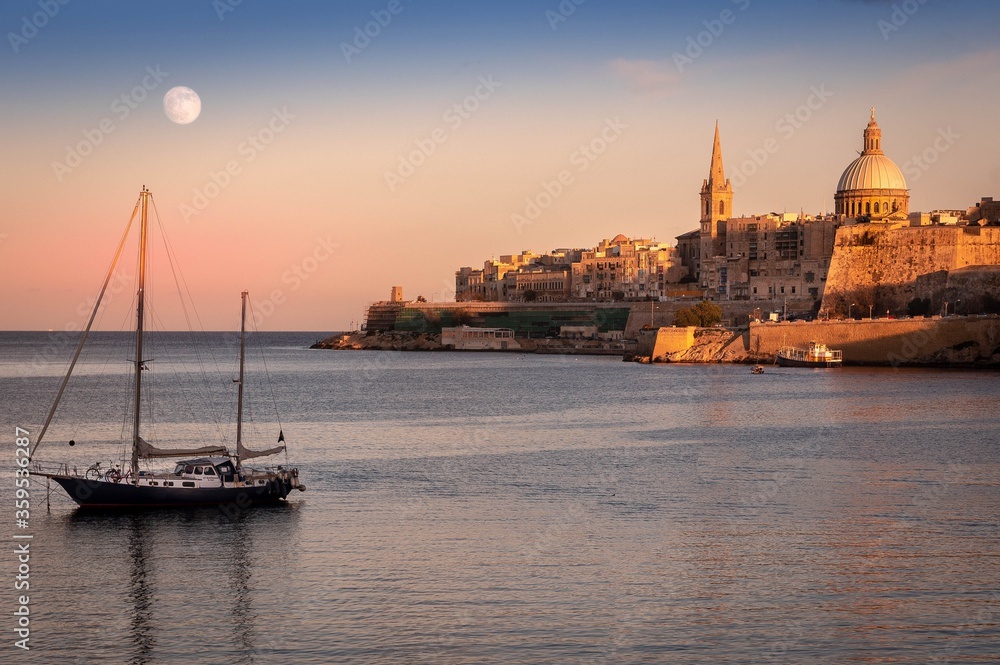 View on Malta walls during sunset with visible moon and a yacht in the foreground. 