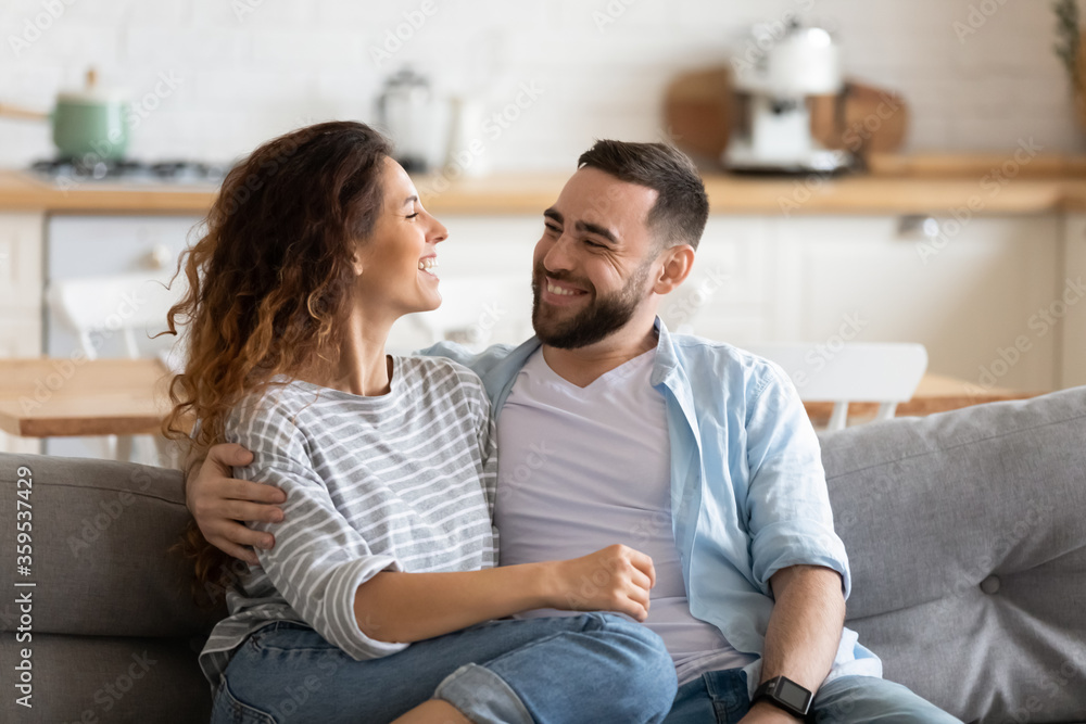 Happy young couple of man and woman embracing look at each other sitting on couch in living room. Attractive smiling husband and wife laughing having fun free time together. Happy young couple of man and woman embracing look at each other sitting on couch in living room. Attractive smiling husband and wife laughing having fun free time together.