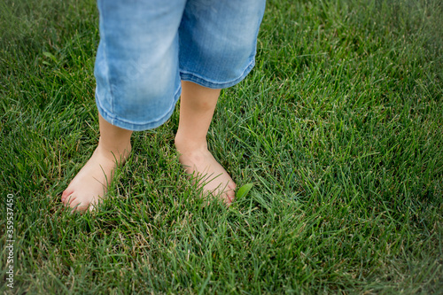 A child stands barefoot on the grass. Only legs in blue denim are visible. Background - bright green grass. Summer. Natural light. Feet took on soft grass.