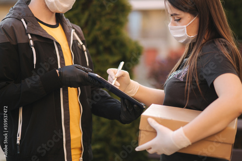 Man from delivery service in medical mask and gloves handing fresh food to young woman customer receiving express delivery from courier at home. Courier with tablet, customer sign in. Female order