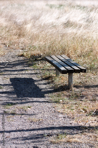 Lone rustic park bench with drop shadow at the edge of a peaceful trail and grassy field