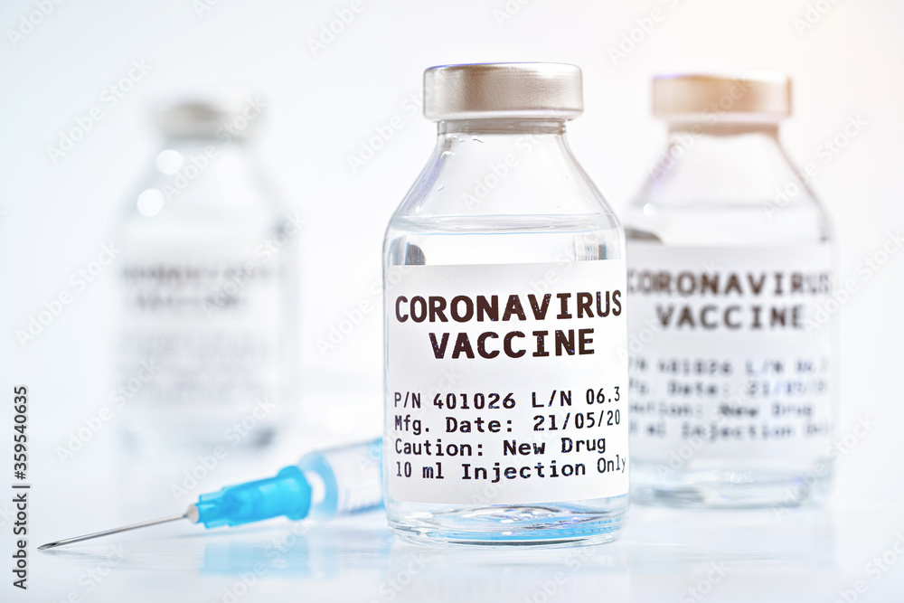 Coronavirus Covid-19 vaccine concept - glass vials with metal caps on white table, blurred hypodermic syringe needle and more bottles in background (label own design - dummy data, not real product) Coronavirus Covid-19 vaccine concept - glass vials with metal caps on white table, blurred hypodermic syringe needle and more bottles in background (label own design - dummy data, not real product)
