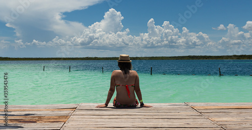 Attractive young female sitting on the border of a pier with the blue water of the Mexican cenotes in the background on a warm summer´s morning  © Enrique