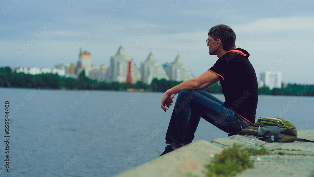 A man with glasses near the river. Portrait of a man in glasses and casual clothes, sitting by the river on a cloudy day