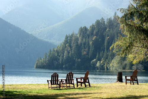 Adirondack chairs in the grass on the side of a lake overlooking pine trees and mountains photo