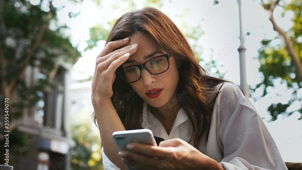 Business woman in glasses, white shirt. She feeling disappointed while reading news using smartphone, sitting in outdoor cafe. Close up, slow motion Business woman in glasses, white shirt. She feeling disappointed while reading news using smartphone, sitting in outdoor cafe. Close up, slow motion