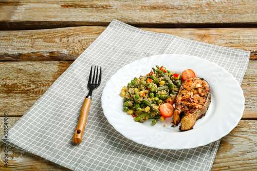 On a linen napkin is a white plate with baked trout steak and a side dish of quinoa with vegetables and a fork next to it.