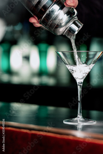 Bartender hand pours vodka or gin from a shaker into a martini glass waiting to be made at a bar counter.