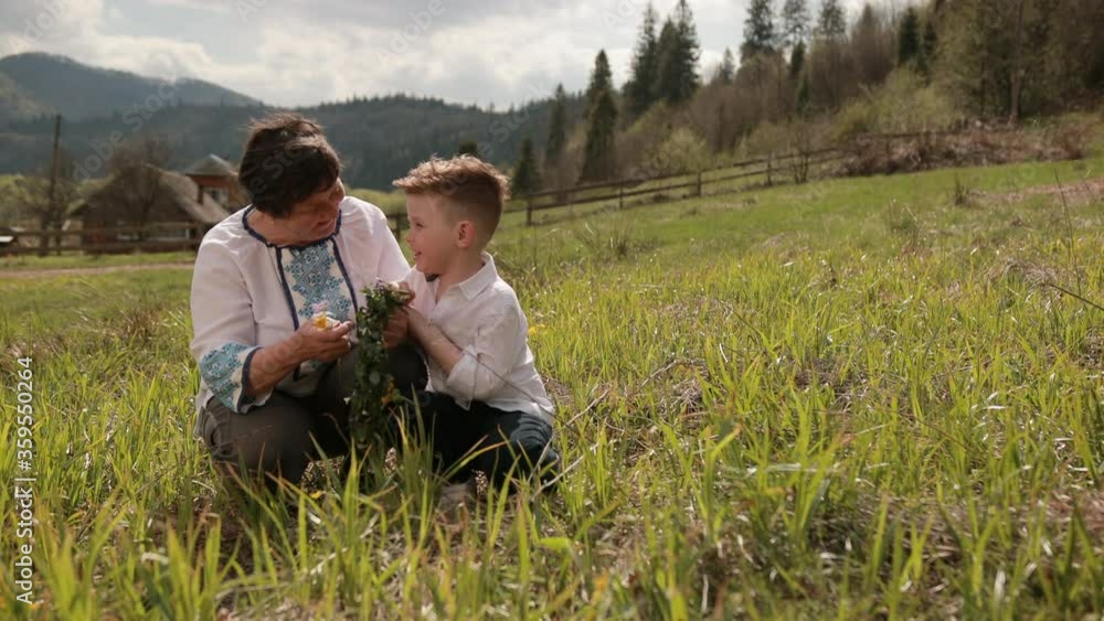 Grandmother makes wreath of wild flowers with her young grandson. Who wears traditional costume embroidery shirt, family making homemade, I love my gran. Relax outside on green grass, behind mountain Grandmother makes wreath of wild flowers with her young grandson. Who wears traditional costume embroidery shirt, family making homemade, I love my gran. Relax outside on green grass, behind mountain