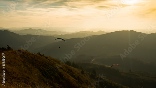 Paragliding concept, Beauty nature mountain landscape. Paraglider silhouette flying over misty mountain valley in beautiful warm sunset.