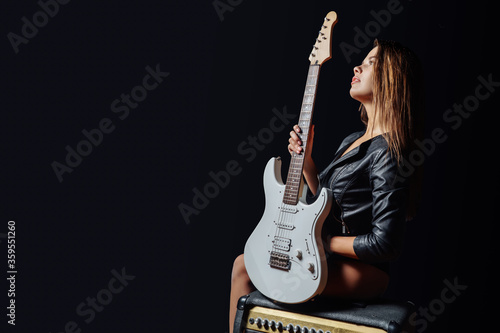 Sexy woman in leather jacket and gloves sitting on a guitar cabinet with an electric guitar