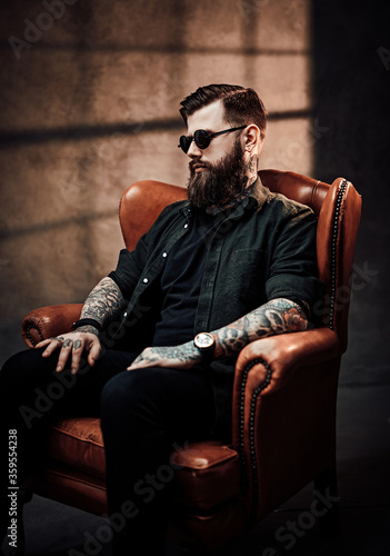 Cool bearded young man sitting on a vintage chair in a dark studio with smoke and window silhouette in the background