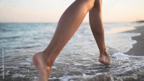 Slim female legs and feet walking along sea water waves on sandy beach. Pretty woman walks at seaside surf. Splashes of water and foam in 120 fps slow motion. Girl after bathing in ocean go on shore. photo