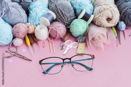 Women's hobby. Multicolored skeins, needles, hooks, glasses on the pink background. Concept of sewing, crochet and knitting.