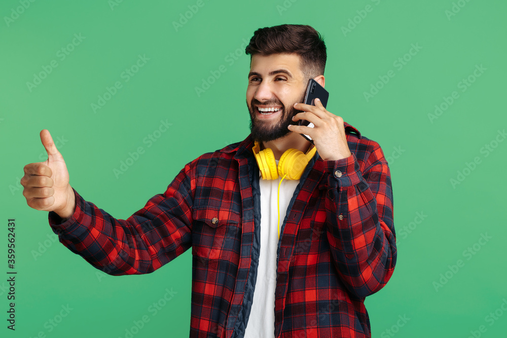 Trendy bearded hipster young man talk on smartphone and show thumb up over green background. Trendy bearded hipster young man talk on smartphone and show thumb up over green background.