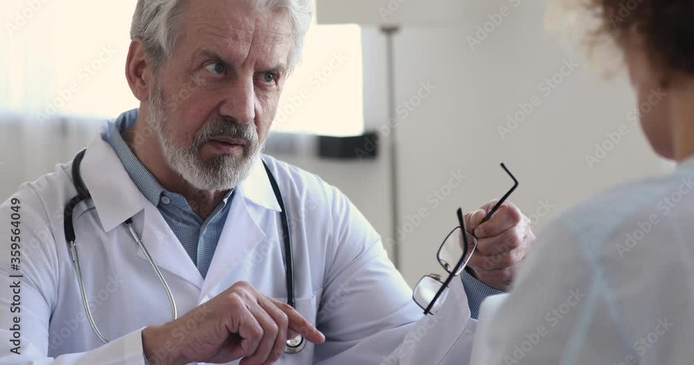Head shot serious middle aged senior hoary male doctor taking off glasses, looking at female patient, explaining treatment or giving professional healthcare advice to young woman at checkup meeting. Head shot serious middle aged senior hoary male doctor taking off glasses, looking at female patient, explaining treatment or giving professional healthcare advice to young woman at checkup meeting.