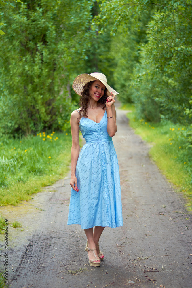 beautiful girl in a straw hat and blue dress on the road in the Park.curly brunette with a smile on her face