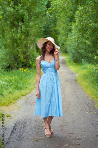 beautiful girl in a straw hat and blue dress on the road in the Park.curly brunette with a smile on her face © евгений ставников