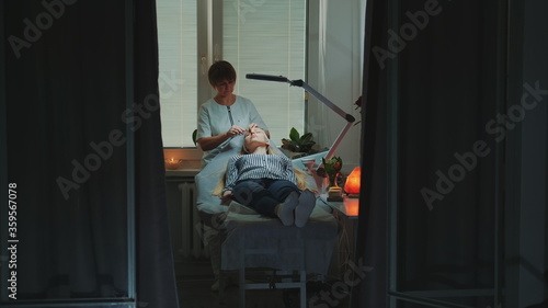 Dolly shot of Beautician making relaxing facial massage with jade roller on woman's face at beauty salon with dim light around and pleasant atmosphere. © art24pro