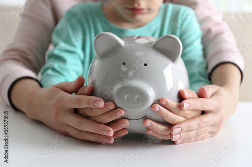Mother and son with piggy bank at white table indoors, closeup