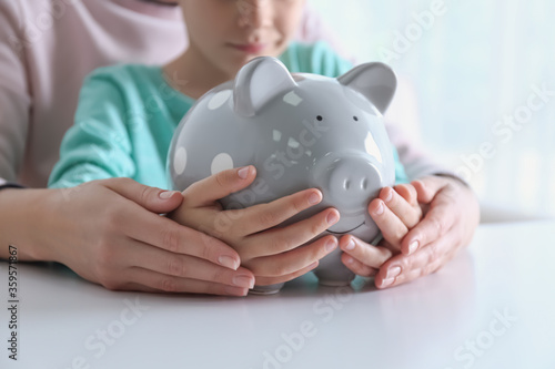 Mother and son with piggy bank at white table indoors, closeup