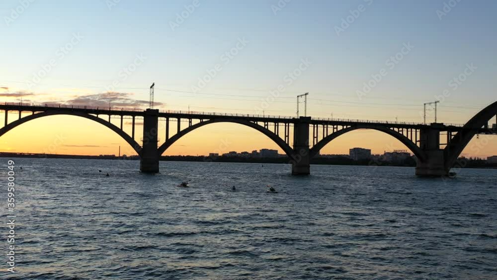 Group of kayakers floats along wide river at sunset under arched bridge. active leisure in city. sport at sunset on background of orange sun. Kayaks dissect surface of river against backdrop of city Group of kayakers floats along wide river at sunset under arched bridge. active leisure in city. sport at sunset on background of orange sun. Kayaks dissect surface of river against backdrop of city