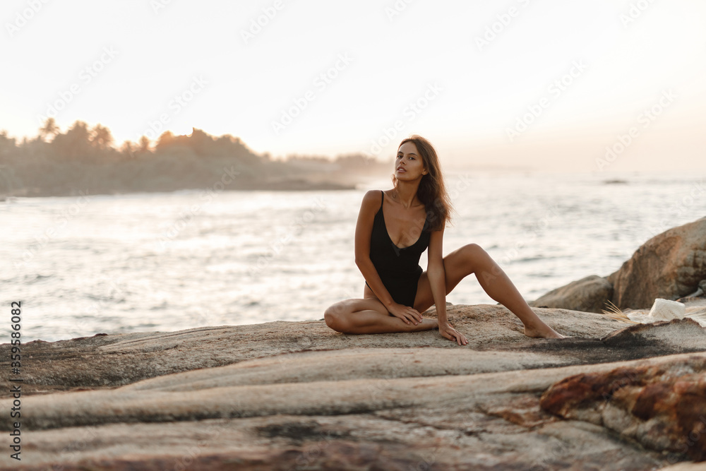 Attractive young girl with long hair is posing to the camera on rocky beach. She wears black bikini and look at the camera