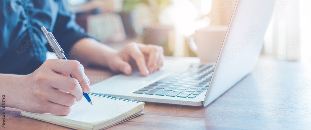 Business woman is writing on a notebook with a pen and using a laptop to work in the office.