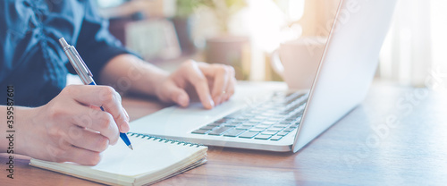 Business woman is writing on a notebook with a pen and using a laptop to work in the office.