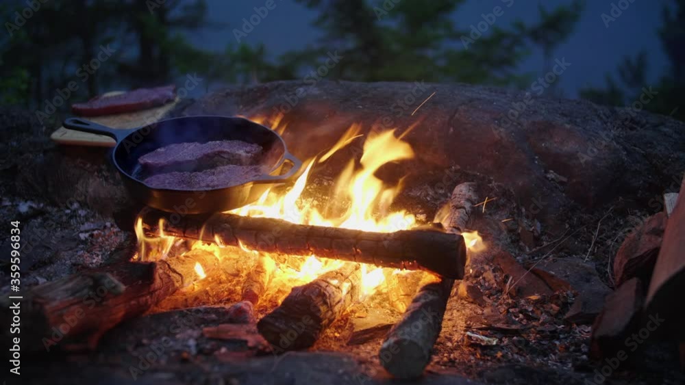 Outdoor cooking over a campfire, grilling steak in cast iron skillet at night Outdoor cooking over a campfire, grilling steak in cast iron skillet at night