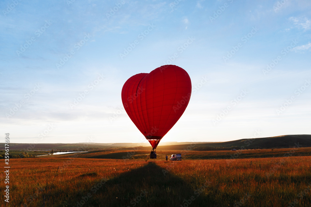Fototapeta premium hot Red air balloon heart shape flying into sunset over valley landing or taking off
