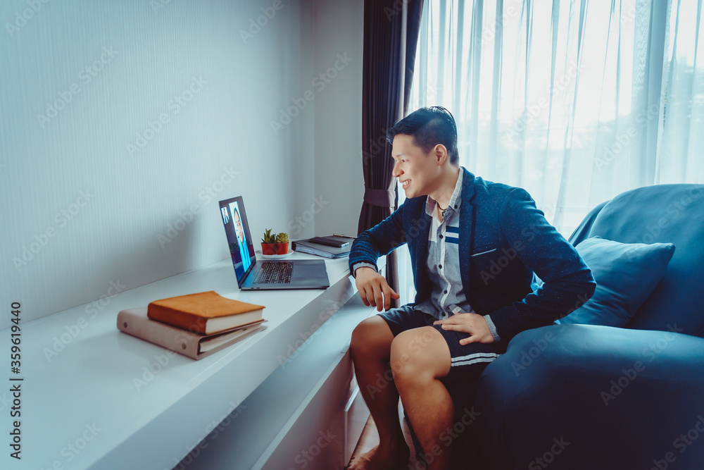Young Asian Businessman wear Formal Suit with Short Pants Having Video Conference Meeting with Business Partner on Sofa at Home in Living Room during Quarantine Period.
