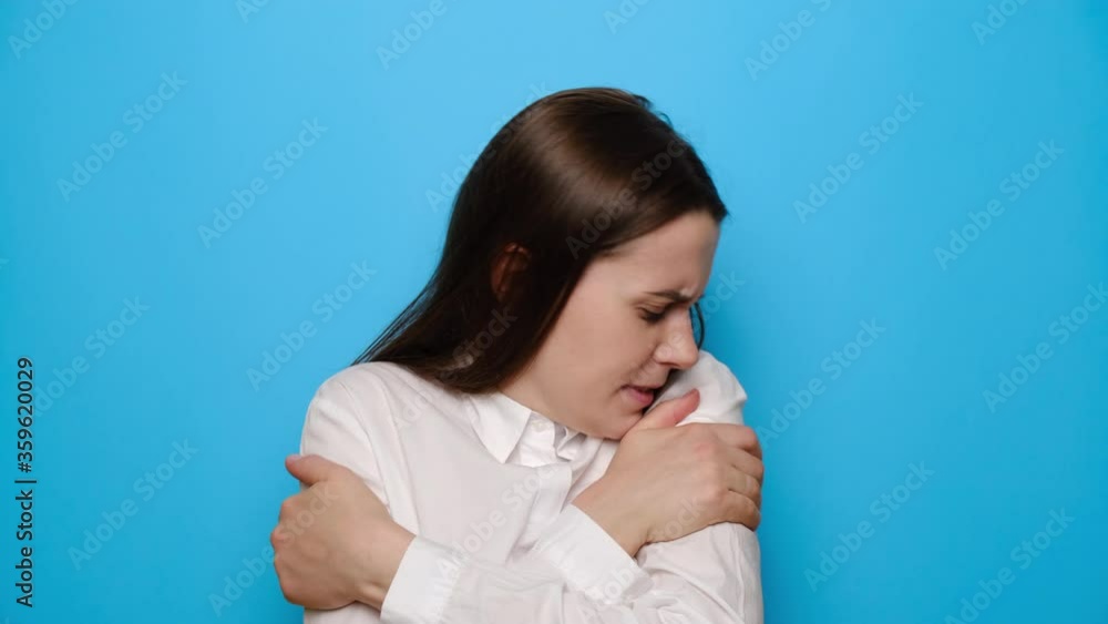 Brunette girl freezes in light clothing warms up by hugging herself, seasonal changes low temperature heating problem, looks at camera, chills feeling of coldness concept, isolated on blue background Brunette girl freezes in light clothing warms up by hugging herself, seasonal changes low temperature heating problem, looks at camera, chills feeling of coldness concept, isolated on blue background