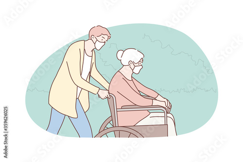 Volunteering, medicine, coronavirus, disability, healthcare concept. Young man volunteer in medical face mask pushing wheelchair old woman granny pensioner. Social support and helping senior citizens.